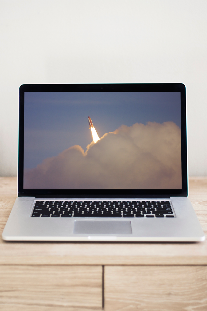 An open Macbook sits on a desk. The screen is an image of a rocket launching through clouds.