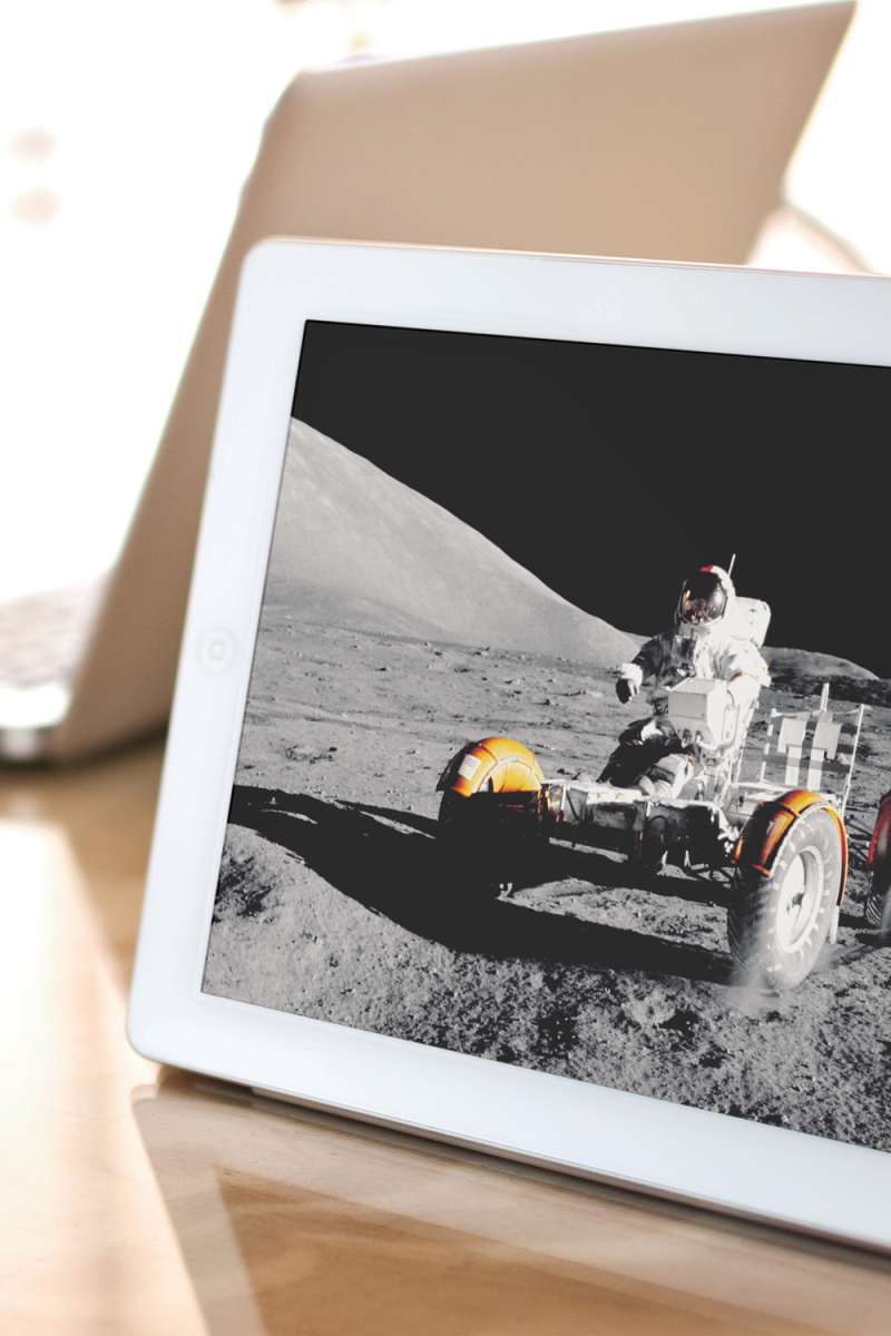 An iPad sits on a coffee shop table. On the left is a cup and saucer. The image on the iPad is an astronaut on a lunar buggy.