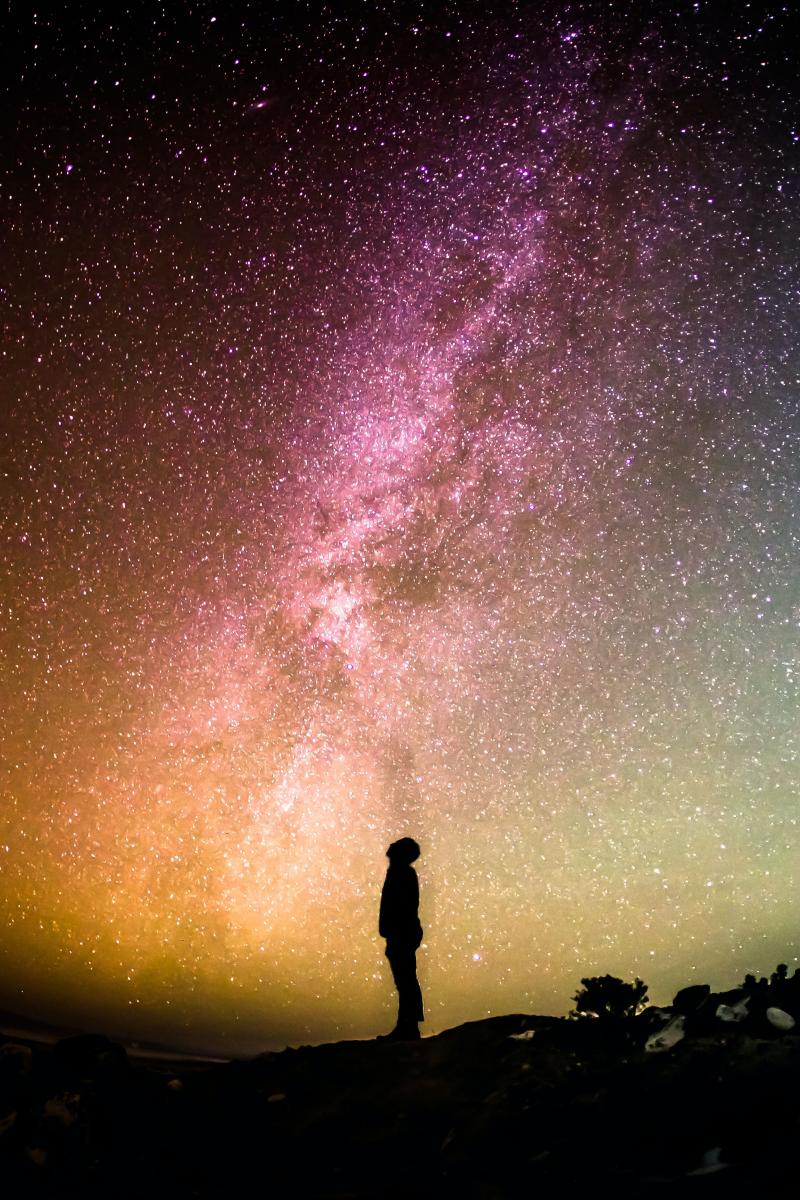 A person stands under a starry sky that is shades of orange, pink and green.