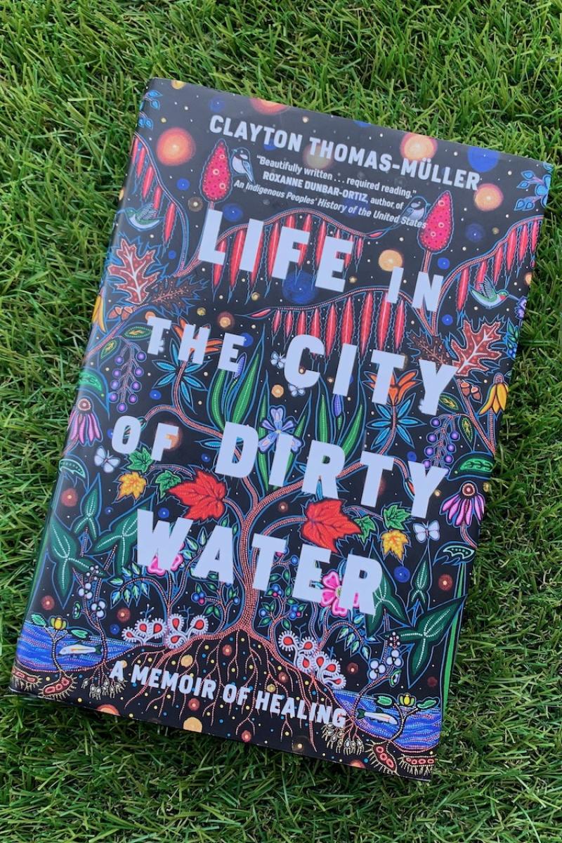 A photo of the book "Life in the City of Dirty Water" by Clayton Thomas-Müller. It is lying on a grass-looking rug.