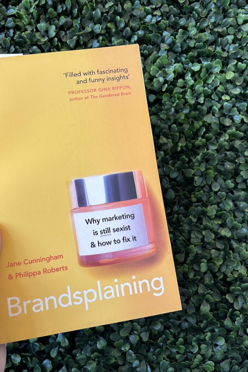 A photo of the book Brandsplaining is held up against a wall of fake green foliage.