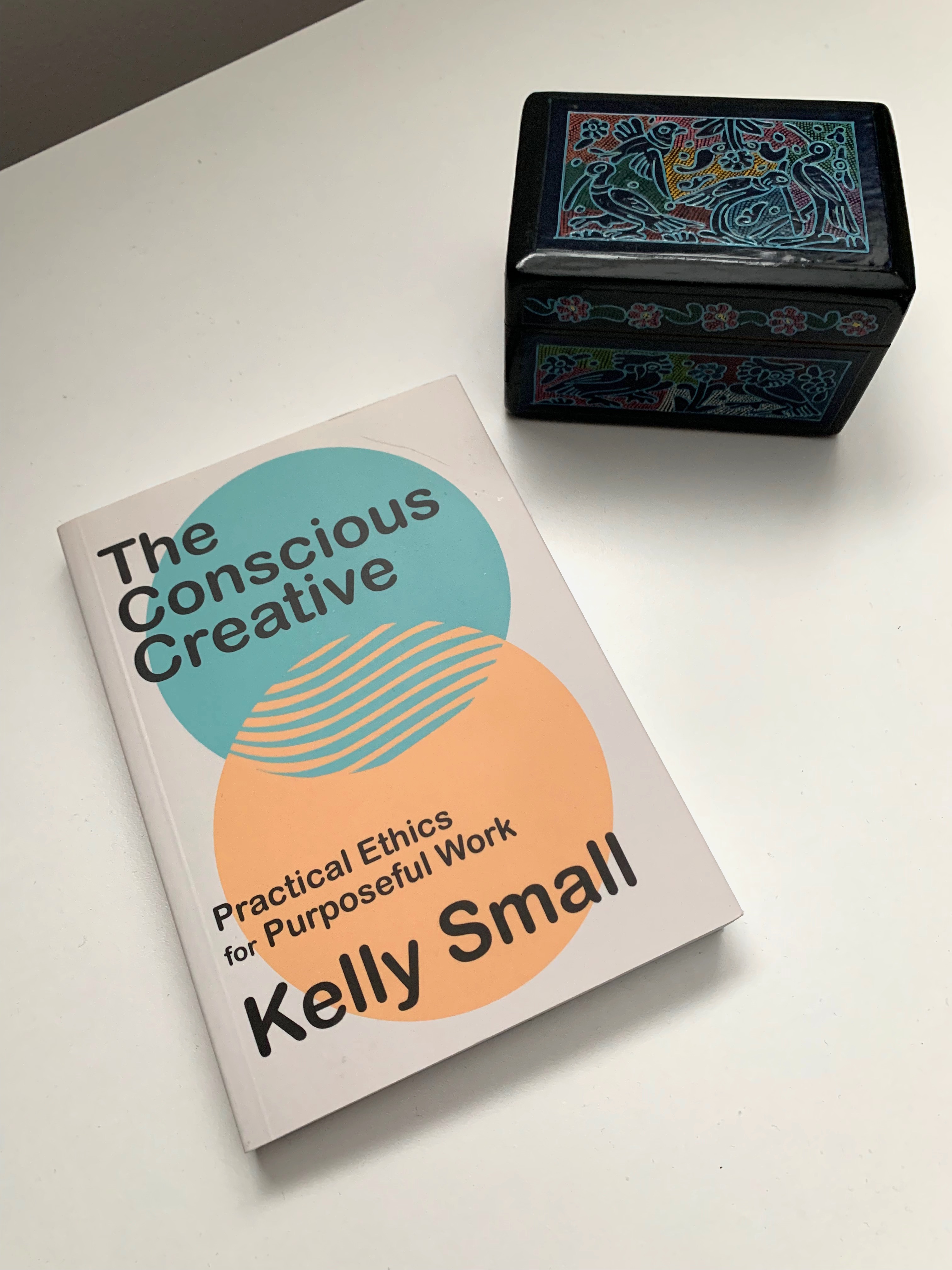 The book "The Conscious Creative" sits on a white desk to the left of a hand made and colourfully painted wood box from Mexico.
