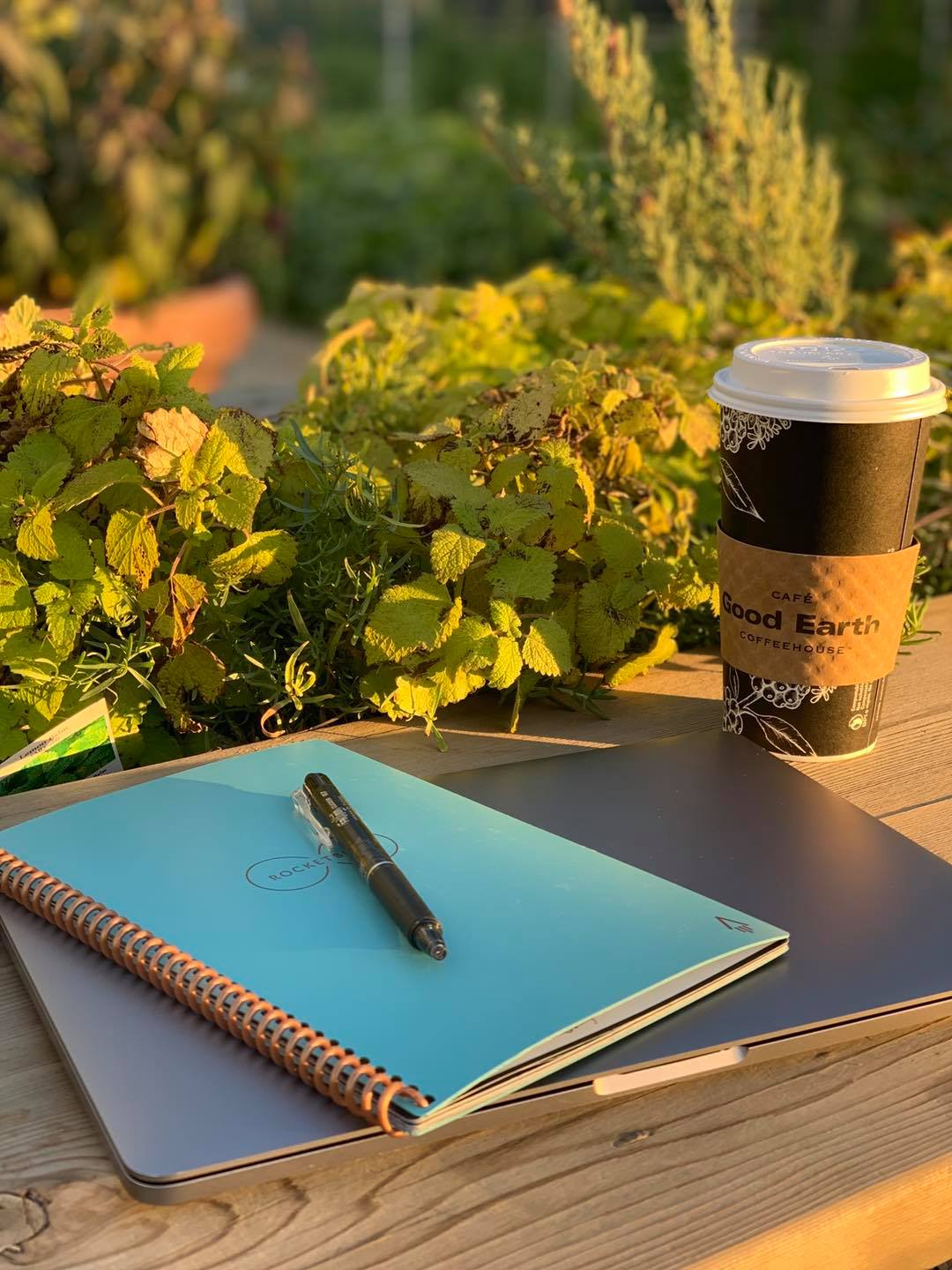 A laptop with a notebook and pen sit atop a picnic table. On the right is a coffee cup. There is greenery in the background.