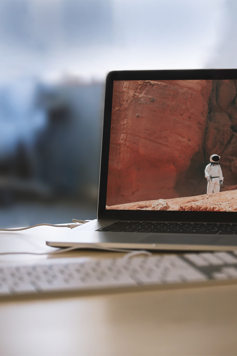 An open Macbook and keyboard are on a desk. The Macbook screen is a photo of an astraunaut on a moon-like surface.
